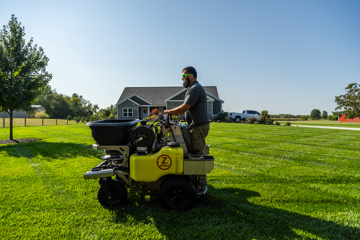 Technician on a ride-on spreader on a large lawn, a gray home in the background