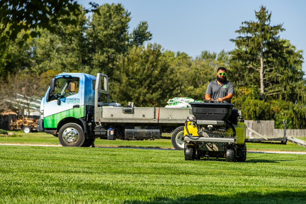 Technician on a ride-on spreader with a Shades of Green truck parked behind