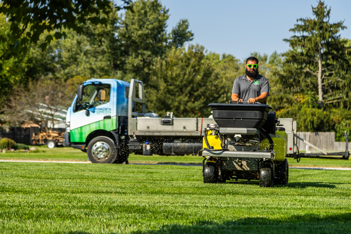 Technician driving a ride-on spreader toward the camera, Shades of Green truck behind