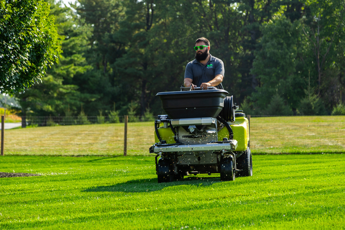 Technician in green sunglasses on a ride-on spreader crossing a striped lawn