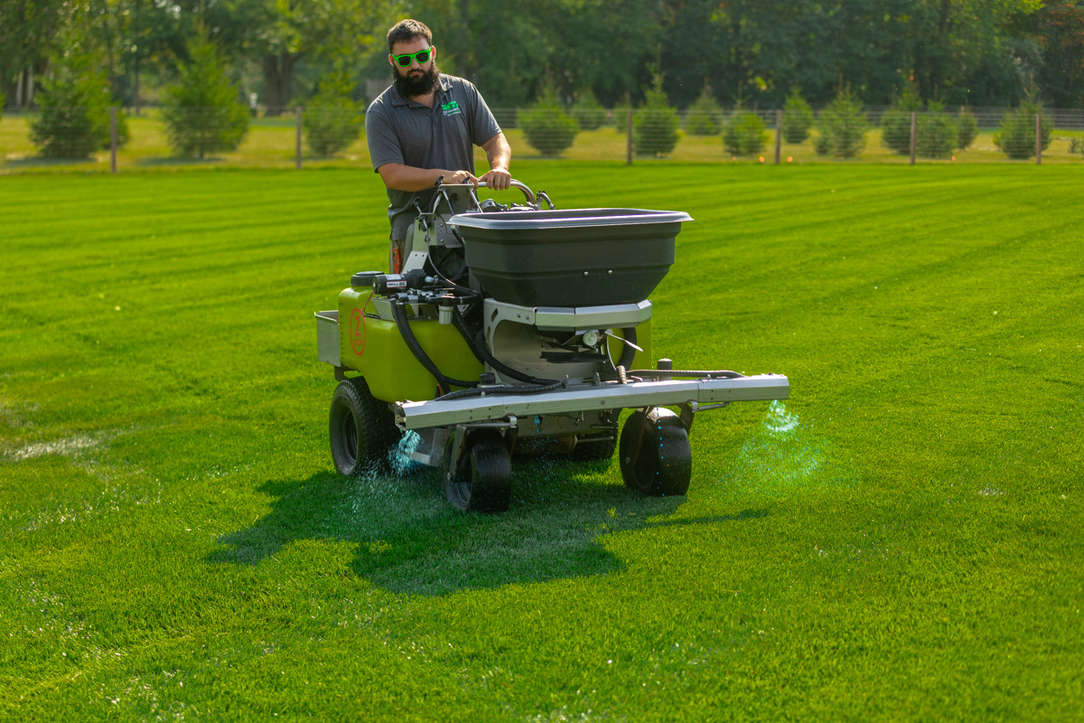 Technician spraying a liquid treatment from a ride-on machine across a striped lawn