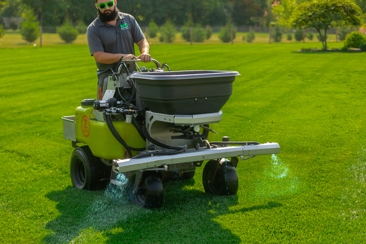 Ride-on machine's boom spraying a blue-tinted liquid treatment on a striped lawn